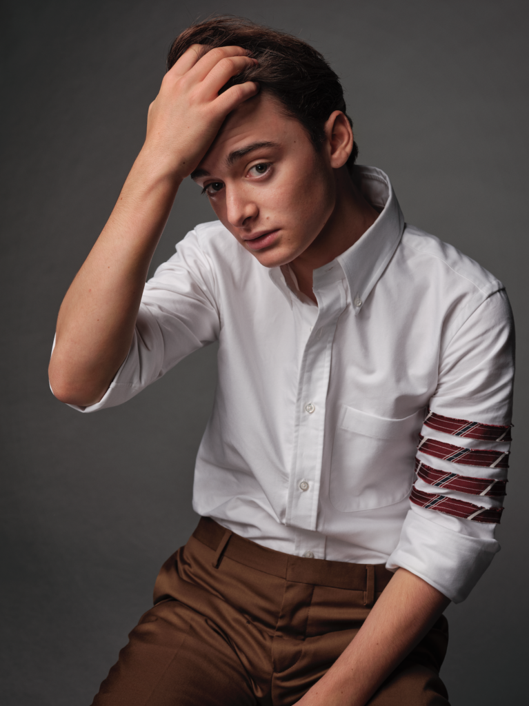 Noah Schnapp puts his fingers in his hair and wears a white collared shirt in a press shot
