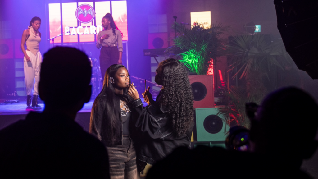 A camera crew stands around Bellah with a stage behind her while she has her makeup done. Two dancers stand onstage behind her with the BACARDÍ logo behind them