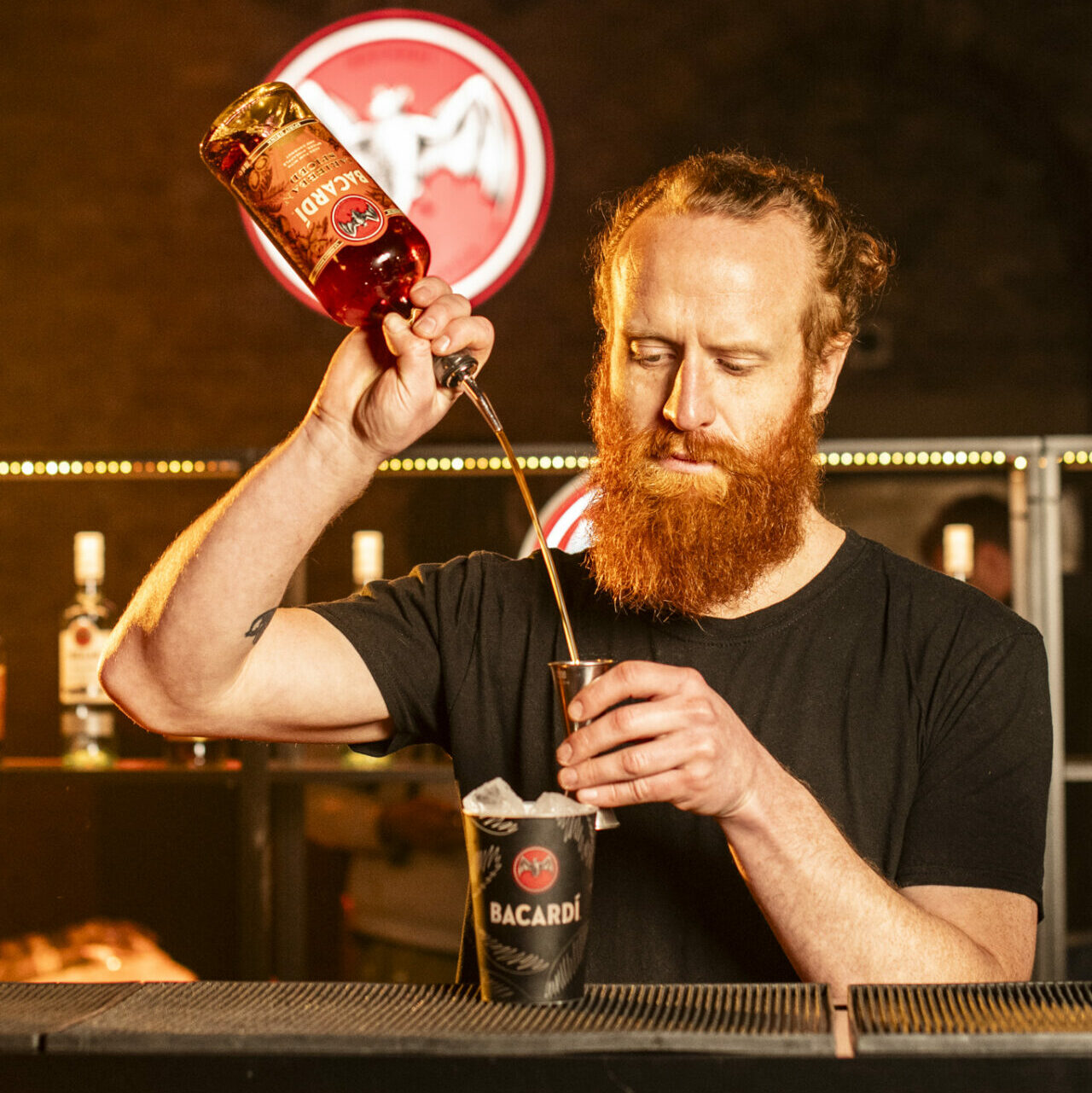 A white man with red hair and beard pours  Bacardí into a glass while standing at a bar