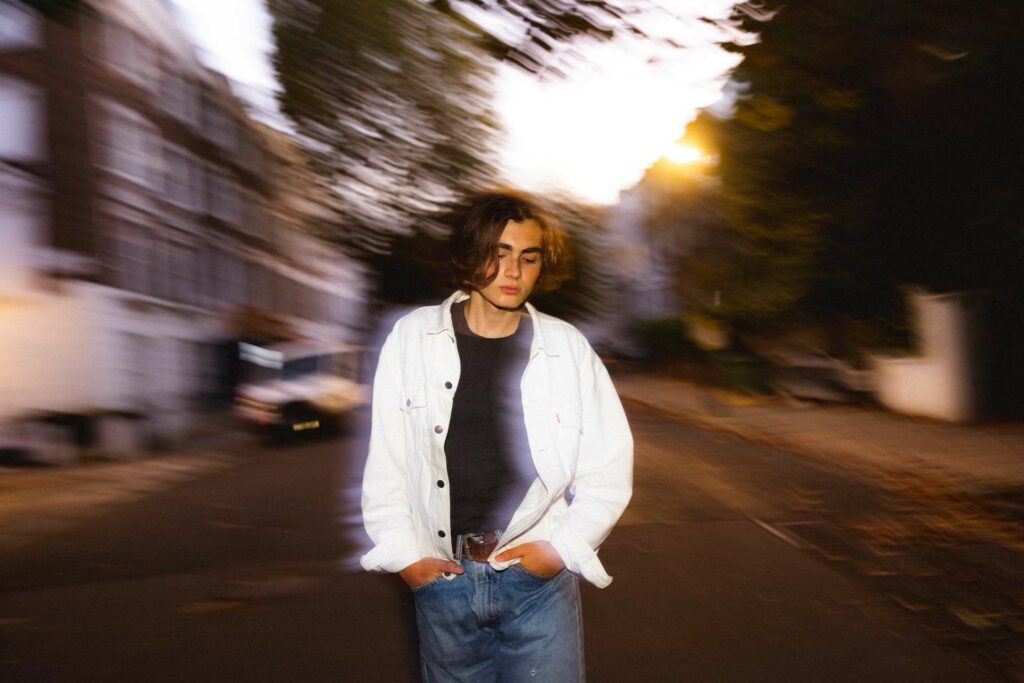 A man wearing a white jacket and blue jeans walking down a street in London with the background blurred