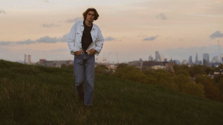 A man wearing a white denim jacket and blue jeans standing on a hillside overlooking London