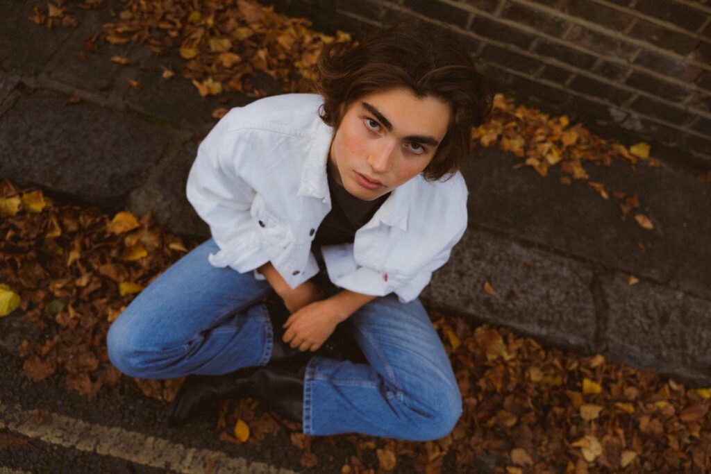 A man sitting on a curb wearing a white jacket and blue jeans looking up into the camera